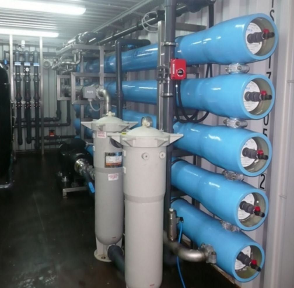 Interior of the reverse-osmosis desalination plant at Elounda Mare, 2024: ranks of pressure vessels with membrane modules, stainless-steel high-pressure piping, control panels.