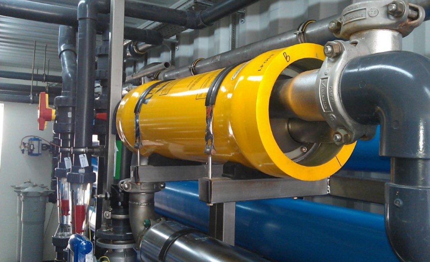 Close detail of the ERI (Energy Recovery Inc.) pressure-exchange unit: a horizontal cylindrical device with stainless-steel manifolds and high-pressure fittings, the technology that recovers brine energy and cuts desalination electricity by approximately ninety percent.
