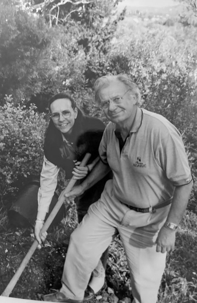 Black-and-white archival photograph of Spyros and Eliana Kokotos together on site, both holding shovels at the ground, working clothes, the early built fabric of the Collection behind them.