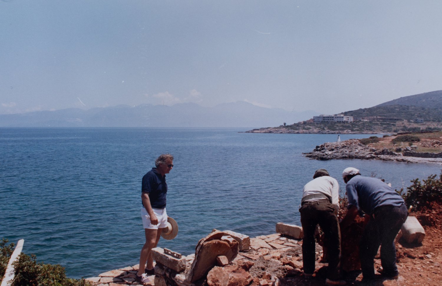 Archival photograph, 1987: Spyros Kokotos at a Mare construction site, in conversation with two masons hand-building a stone wall, the gulf behind.