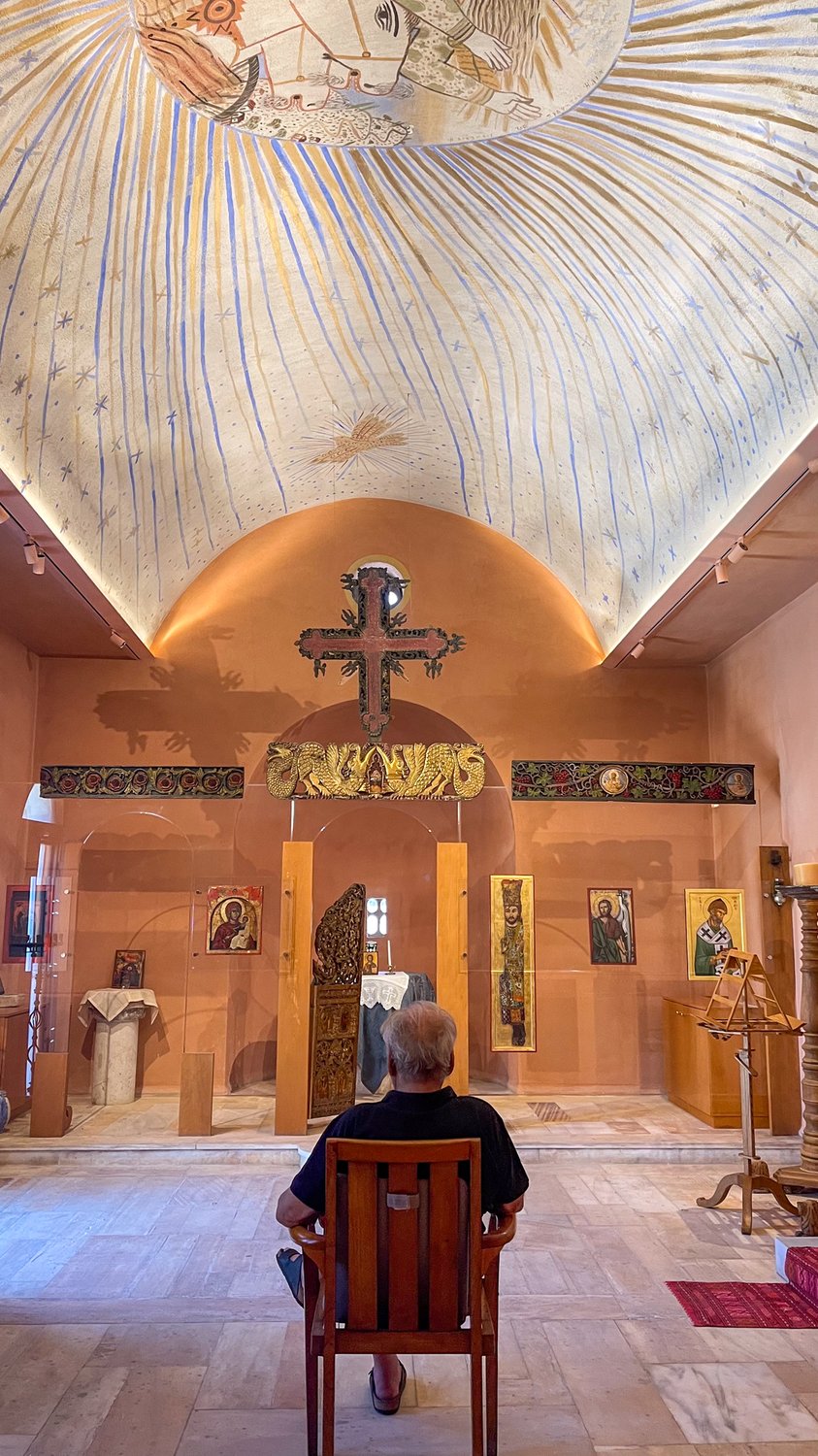 Spyros Kokotos in the Chapel of the Prophet Elias at Porto Elounda, around 2010, standing within his own commission beneath the Fassianos hagiography of the Ascension on the chariot of the Sun.