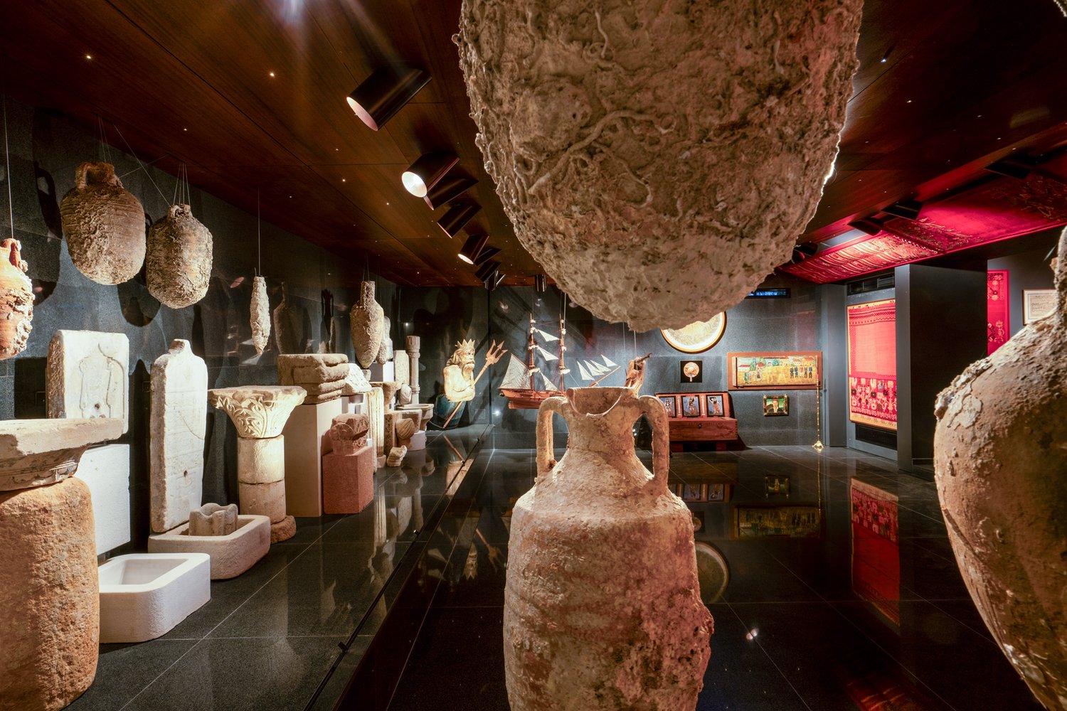 Interior of the Museum of Cretan Folk Art at the Varouhas Estate: the antiquities room, with hanging amphorae and salvaged stone elements against dark stone walls; the polished black floor reflects them.