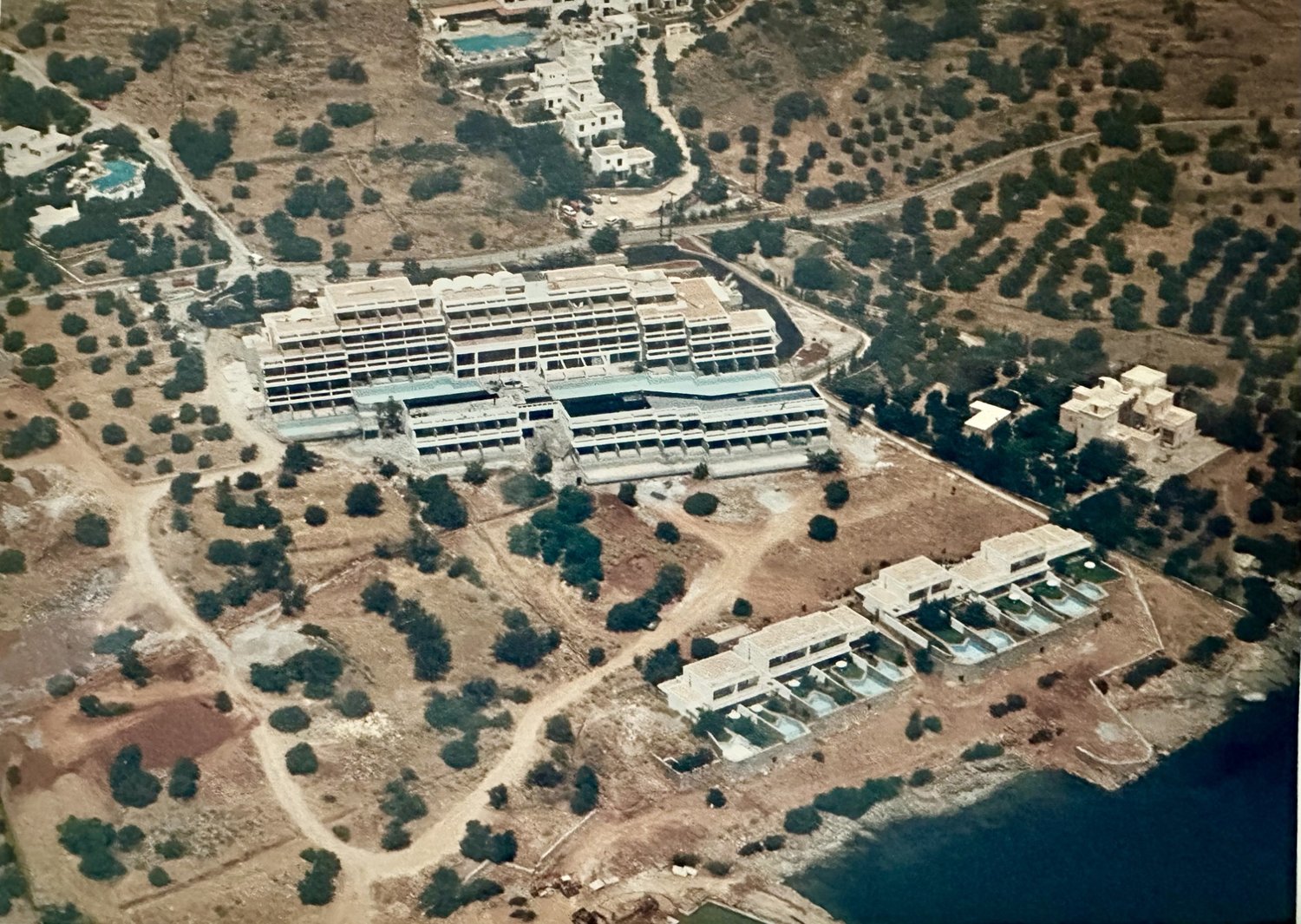 Aerial photograph of Porto Elounda in 1992, its opening year — main hotel block, bungalows with private pools at the water's edge, olive groves around. From the family archive.