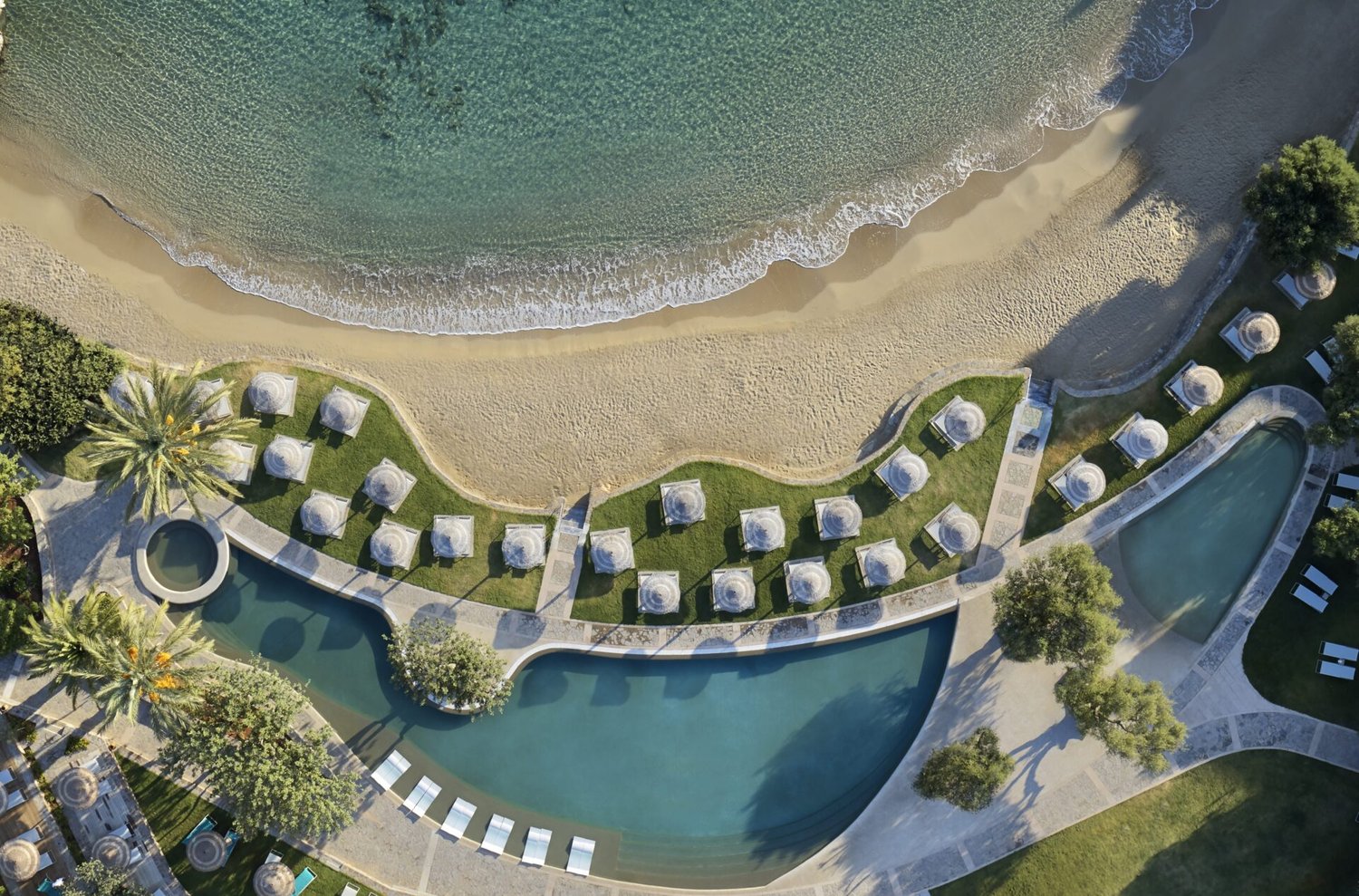 Aerial of Porto Elounda from above: the curved seaside pool meeting the beach, the property opening to the gulf.